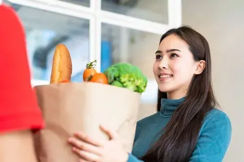 Grocery handoff: a smiling woman receives a paper bag full of fresh produce from another person, utilizing last mile tracking to ensure a friendly exchange or delivery of healthy food.