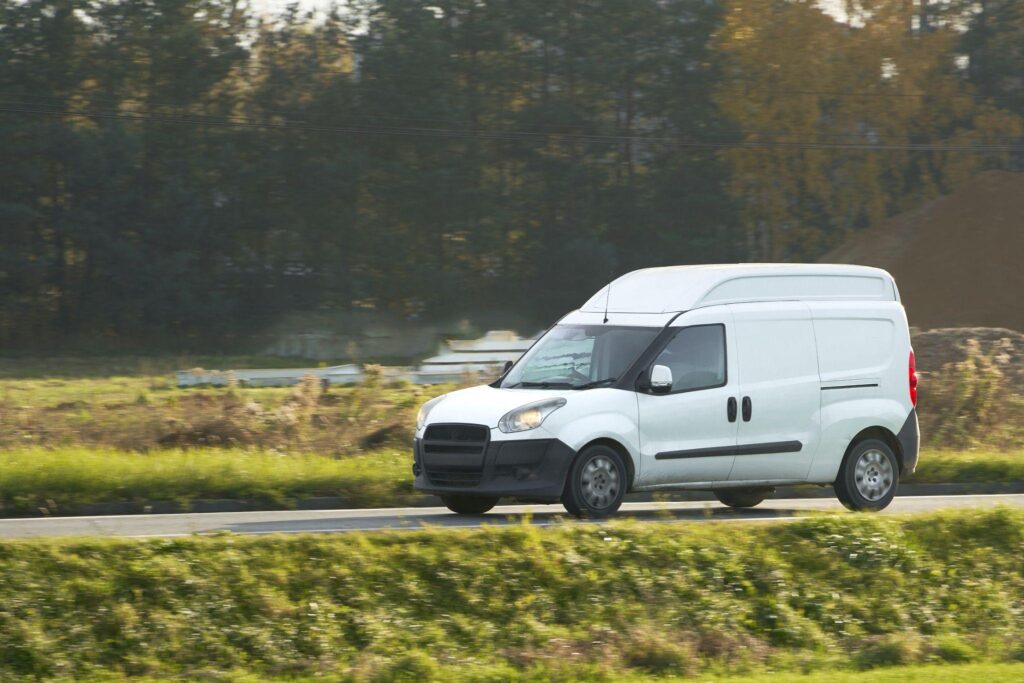 A wide shot of a white delivery van driving along the road above a grass bank