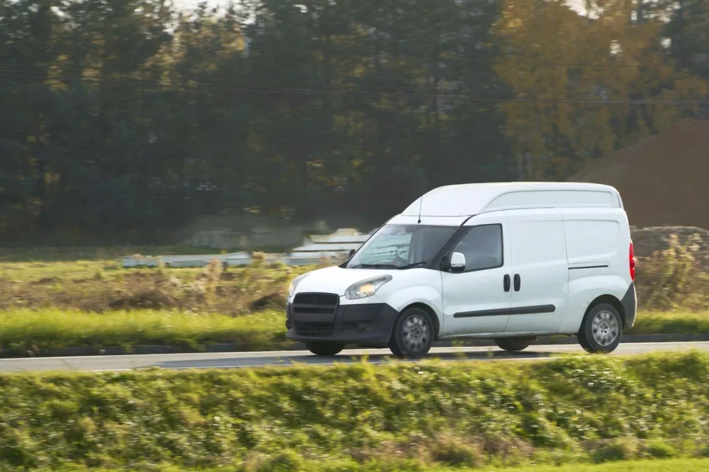 A wide shot of a white delivery van driving along the road above a grass bank