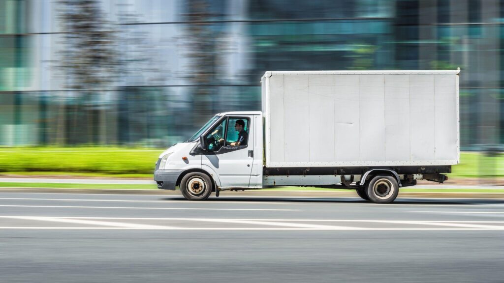 Compact urban box truck passing modern buildings, depicting delivery route optimization within dense city corridors.