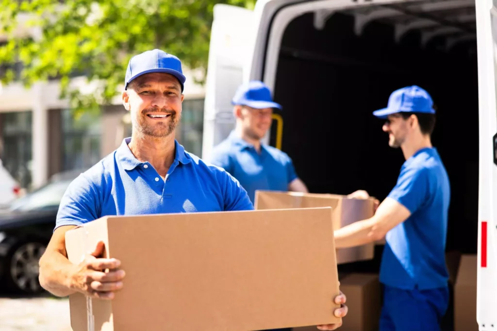 Worker smiling while carrying a cardboard box near a parked delivery van.