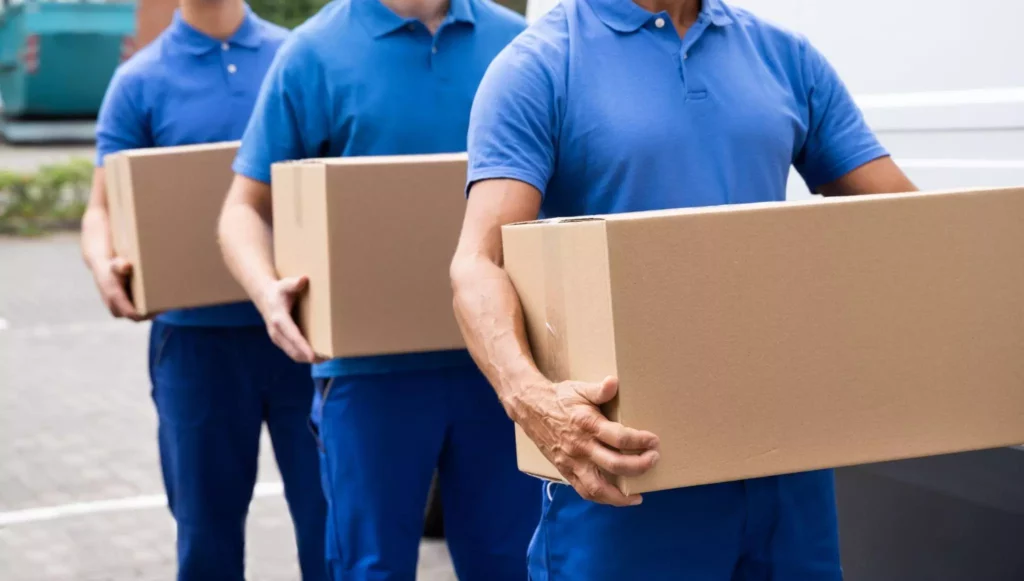 Three delivery workers in blue shirts walking in a line while holding cardboard boxes