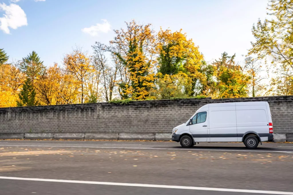 White delivery van driving along a roadside with autumn trees in the background