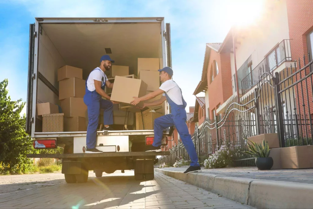 Two workers passing a large cardboard box to each other at the back of a moving truck parked along a residential street