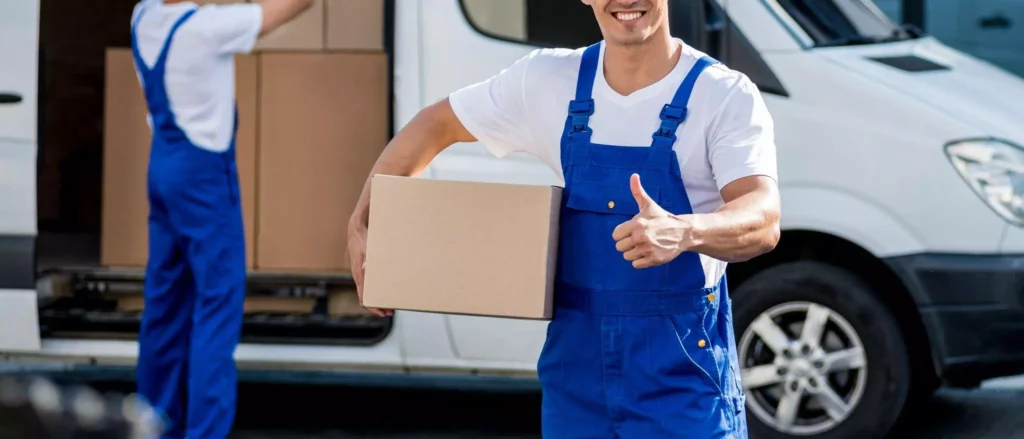 Smiling worker in blue overalls holding a cardboard box and giving a thumbs up near a delivery van