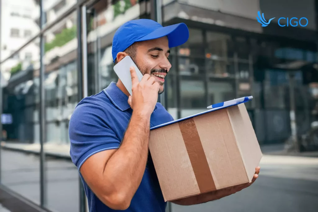 Smiling delivery driver holding a parcel and talking on the phone, illustrating customer communication tools integrated with live delivery status updates.