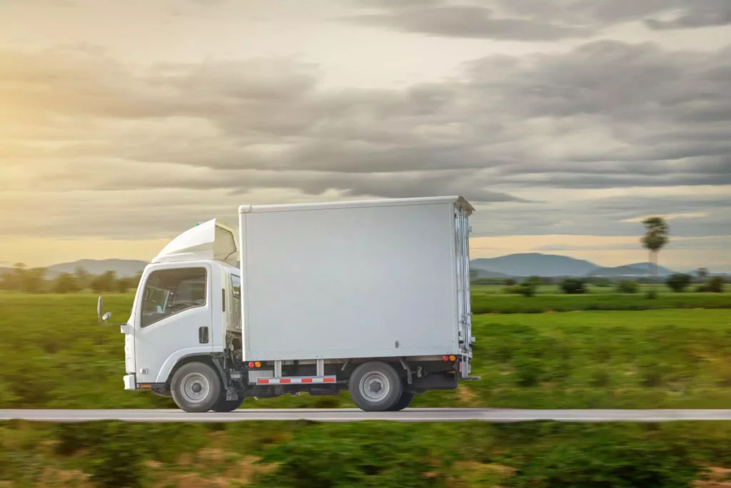 White box truck driving along a rural road with open fields in the background.