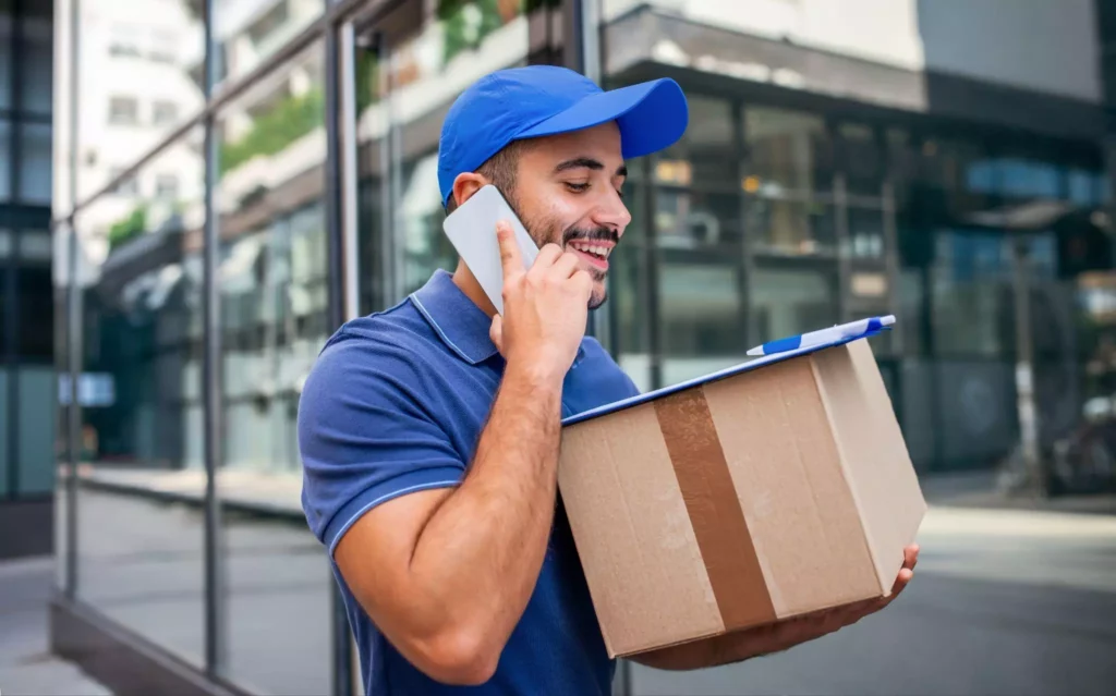 Smiling delivery driver holding a parcel and talking on the phone, illustrating customer communication tools integrated with live delivery status updates.