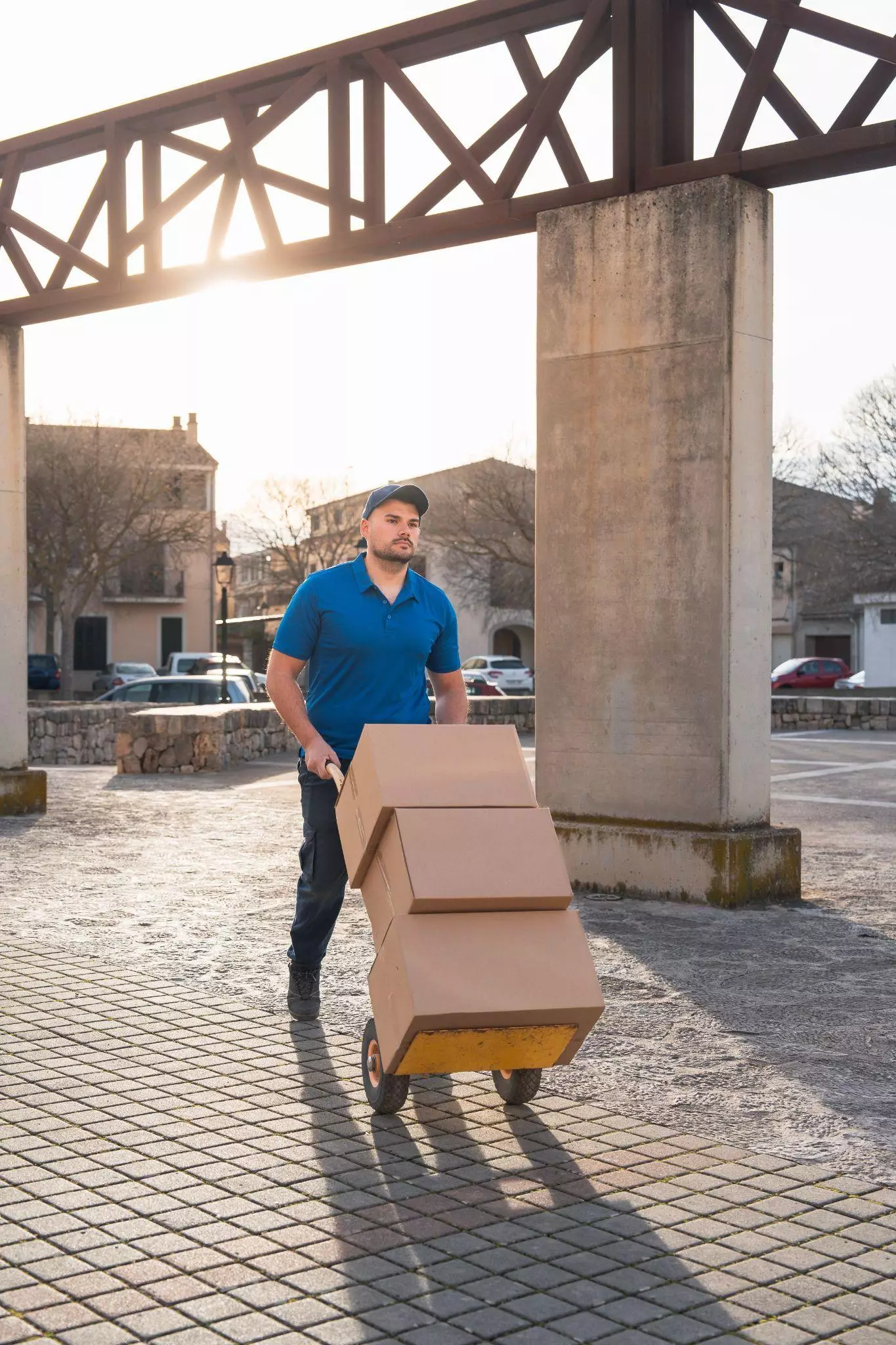 Delivery worker in a blue shirt pushing stacked boxes on a hand truck in an outdoor area.